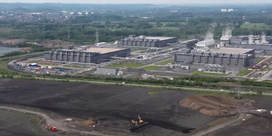 Dronebeelden tonen een industrieel gebouwencomplex aan de rand van een veld vol zand en bouwvoertuigen.