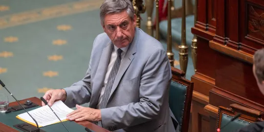 Jan Jambon, in een lichtgrijs kostuum, zit achter het spreekgestoelte in de Kamer. 