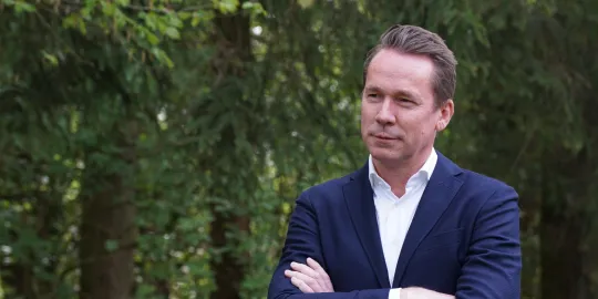Jo Brouns in een donkerblauw kostuum en met de handen gekruist voor een aantal bomen in het Hallerbos.