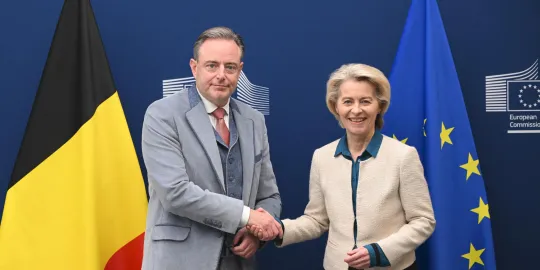 Bart De Wever en Ursula von der Leyen schudden elkaar de hand voor een Belgische en Europese vlag.