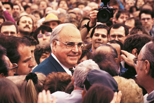 Helmut Kohl wordt omringd door een grote groep mensen, met ook een fotograaf die een foto probeert te maken.