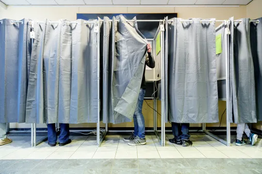 Vijf stemhokjes op een rij, met dichte grijze gordijnen, waar enkel de voeten van de personen te zien zijn. Een persoon stapt het hokje net binnen en trekt de gordijn achter zich dicht.