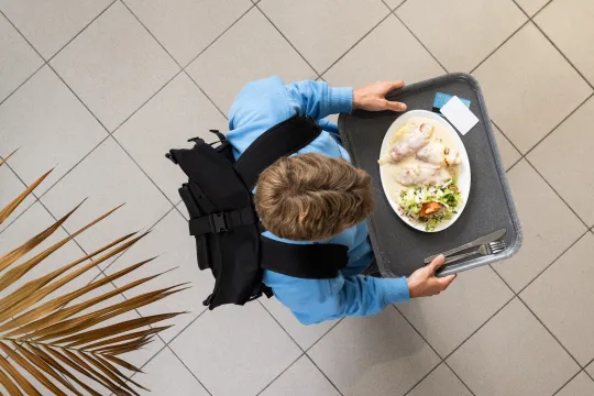 Bovenaanzicht van een student die met een dienblad met daarop een bord vol eten door een studentenrestaurant wandelt.