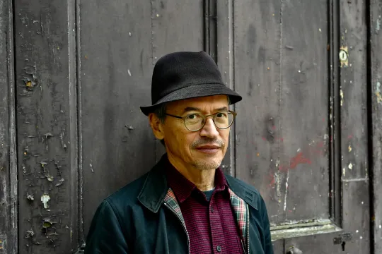 Portret van Joe Sacco, die leunt tegen een grijze, afbladderende deur.