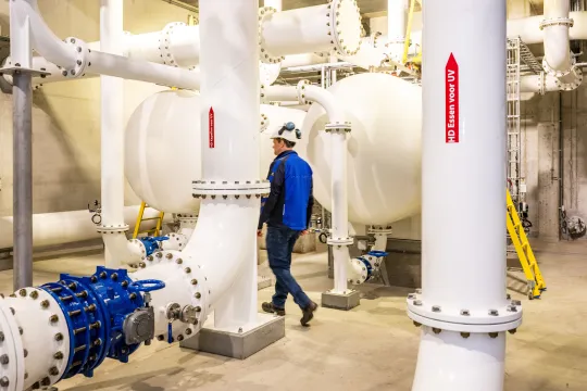 Een persoon met blauwe werkkledij en witte helm loopt door het drinkwaterwinning/productiecentrum van Pidpa in Essen.