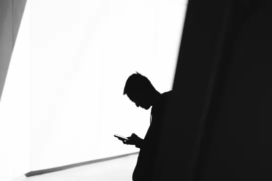 Een persoon kijkt met neergebogen hoofd naar diens smartphone, het beeld is overbelicht en toont de persoon in profiel, in de schadwuw. Rechts is een zwarte voorgrond, links een witte achtergrond, met de persoon ertussen.