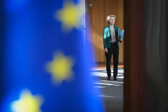 Voorzitter Ursula von der Leyen voor een vergadering van de Europese Commissie in Brussel. Op de voorgrond is een wazige Europese vlag te zien.