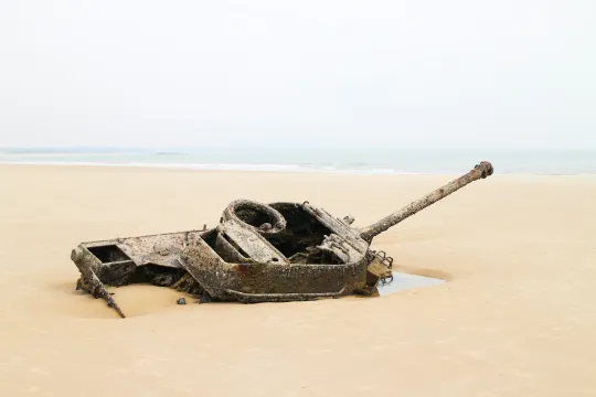 Een tank zakt weg in het zand op een strand, met de zee in de achtergrond.