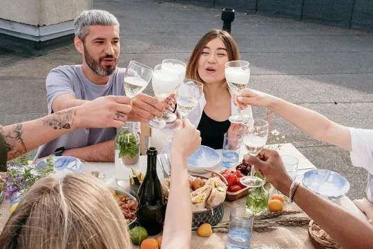 Zes mensen laten hun wijnglazen klinken boven een rijkgevulde tafel met eten, buiten op een dakterras.