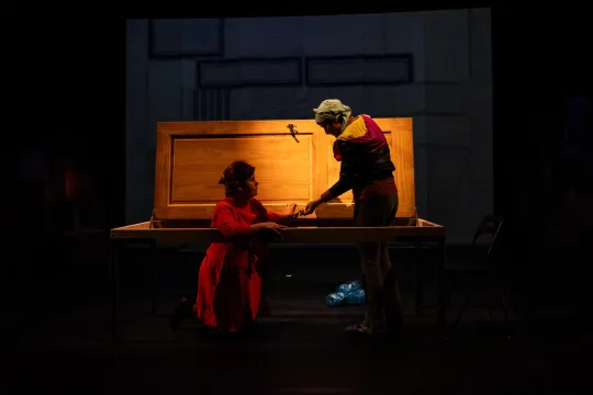 Een vrouw klimt door een horizontale deur op een podium, en wordt de hand gereikt door een andere vrouw