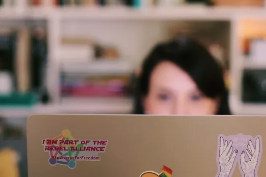 Op de voorgrond is de achterkant van een laptop te zien met een sticker 'I am part of the rebel alliance - #FediverseForFreedom', in de achtergrond het wazige hoofd van een persoon met langer zwart haar die voor een boekenkast zit.