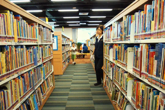 Vrouw en een jong kind lopen langs rekken gevuld met boeken in een bibliotheek. 