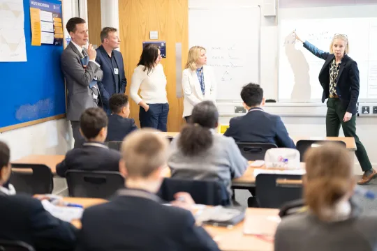 Een rijtje onderwijsexperts staan vooraan links voor een klas, terwijl een leerkracht iets uitlegt om een digitaal bord.