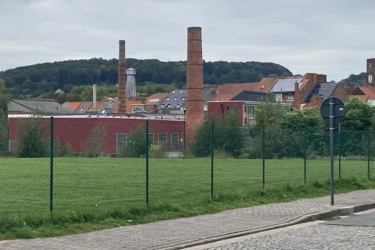 Vanop een verlaten straat kijk je op een leeg grasveld met daarachter een aantal huizen en de schoorsteen van een fabriek. 
