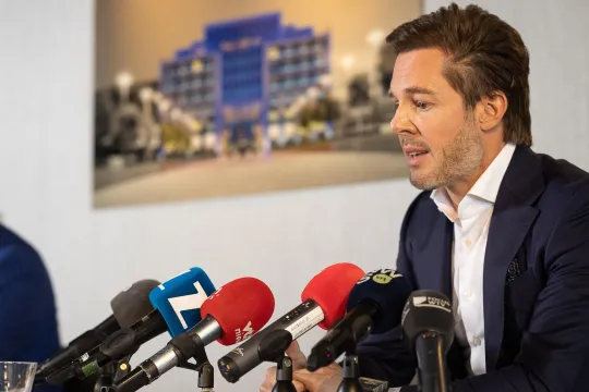 Bart Versluys zit in maatpak voor enkele microfoons tijdens een persconferentie
