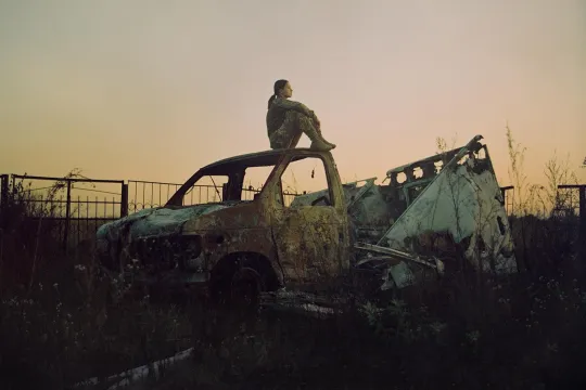 Een vrouw zit in legeruniform op een compleet vernielde truck naar de ondergaande zon te kijken.