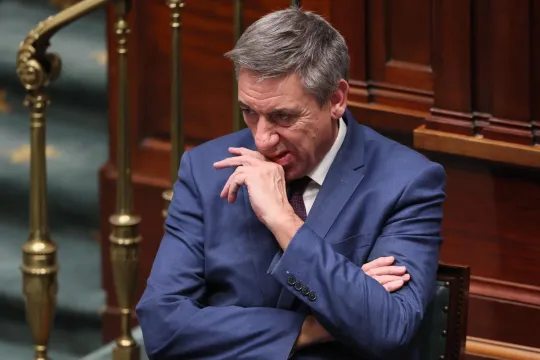 Jan Jambon zit, in blauw pak, in het Federaal Parlement met gekruiste armen en een bedenkelijke blik.