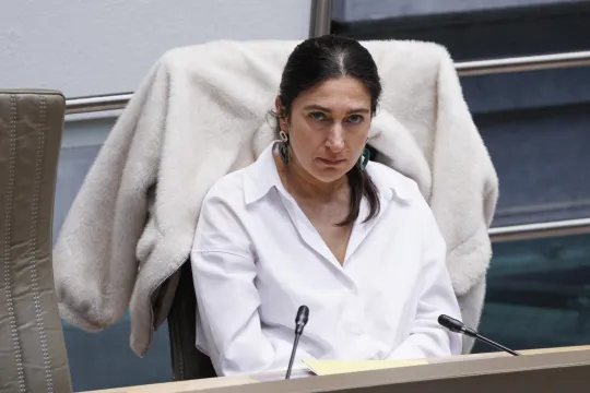 Zuhal Demir, in wit hemd, zit met een verveelde uitdrukking in het Vlaams Parlement, een witte vest over de rugleuning van haar stoel.