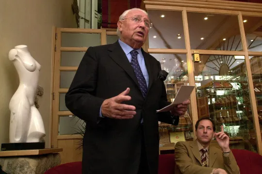 Rudolf Vanmoerkerke staat recht en leest op van een papier, geflankeerd door een journalist van Knack en een buste van een naakte vrouw. 