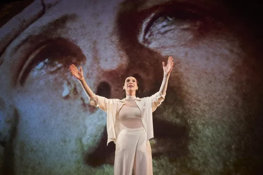 Een vrouw in het wit gekleed heft haar armen op naar een videoprojectie van een schilderij met Maria Magdalena