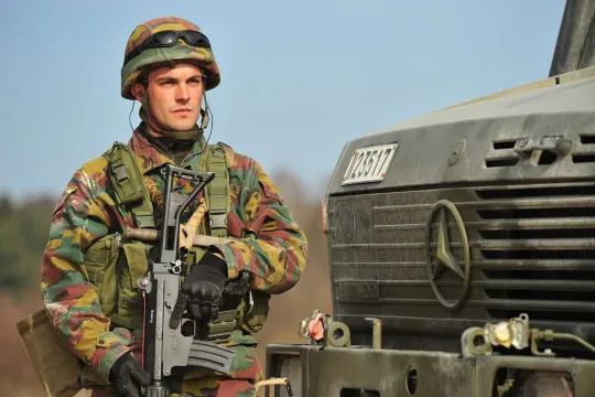 Een soldaat staat in een camouflage-outfit en met wapen naast een militair voertuig. 