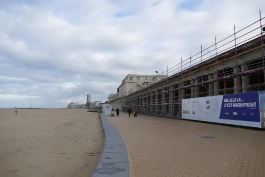 Voetgangers wandelen op de dijk van Oostende voorbij de steigers voor de gaanderijen van het Thermae Palace