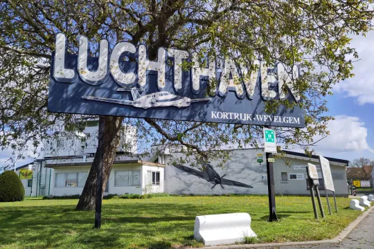 Voor een gebouw, onder een boom, staat een groot bord met in witte letters het woord 'Luchthaven Kortrijk-Wevelgem'.