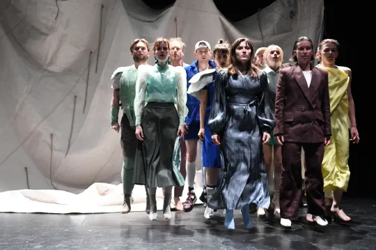 Een groep vrouwen en mannen in sjieke klederdracht staat op het podium, gefotografeerd in het midden van een rechte sprong en tegen een achtergrond van een wit, gedrapeerd laken.