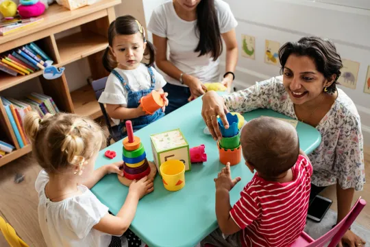 Twee kinderopvang-begeleidsters zitten aan een tafeltje met drie kinderen die met blokken en torens aan het spelen zijn. 