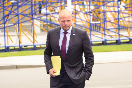 Theo Francken, in een donkergrijs kostuum met een paarse das, wandelt op de camera af. Hij kijkt naar de grond en houdt een papieren map vast. 