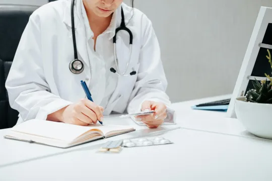 Een dokter in een witte jas zit aan haar bureau, bekijkt medicatie en maakt notities. 