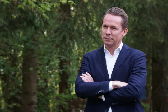 Jo Brouns in een donkerblauw kostuum en met de handen gekruist voor een aantal bomen in het Hallerbos.