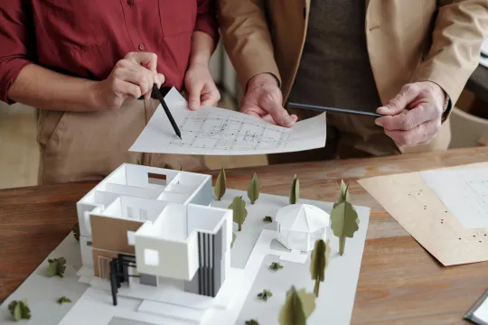 Twee architecten bekijken een maquette van een huis omringd door bomen. Een van hen wijst iets aan op een papier, we zien enkel hun handen.