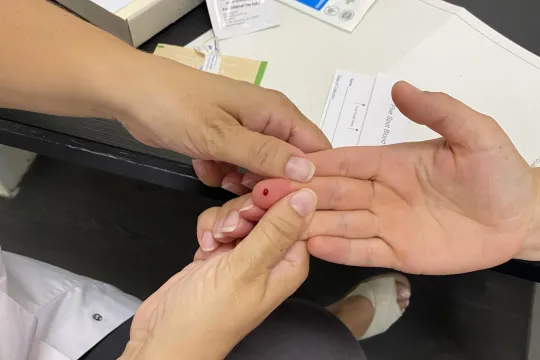 De handen van een verpleger houden de hand vast van een proefpersoon, op wiens vinger een druppeltje bloed te zien is. 