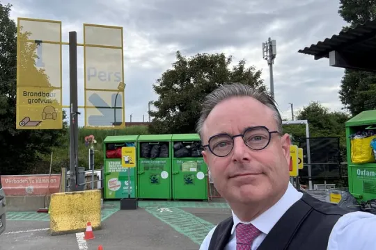 Een selfie van Bart De Wever, in kostuum en met das, waarop achter hem een aantal containers en een banner met de woorden 'pers' te zien is op het containerpark.