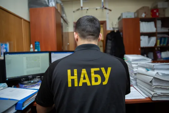Achteraanzicht van een man, met het Russisch 'Nabu' op zijn t-shirt, die op een computer tekst aan het lezen is aan een bureau vol met stapels papier. 