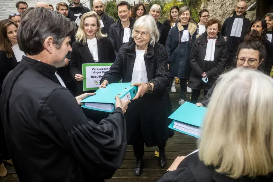 Een groep advocaten in toga staan in een cirkel rond twee advocaten, die een grote dossiermap doorgeven.