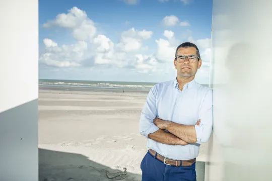 Sander Loones, in een lichtblauw hemd, staat met zijn armen gekruist en kijkt naar de camera. Achter hem de zee.