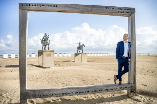 Sander Loones, in een donkerblauw kostuum, staat in een groot houten kader aan zee. 