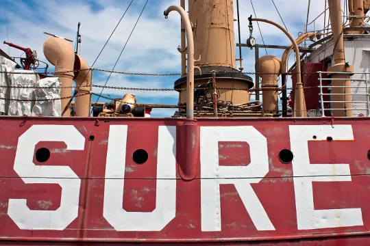 Zijaanzicht van een groot schip met op de zijkant met gigantische letters het woord 'sure' geschilderd. 