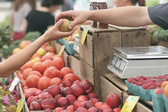 Op een groenten- en fruitkraampje op de markt reikt de ene persoon de andere een appel aan. 