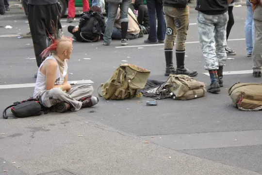 Een punker, met deels geschoren hoofd en een hanenkam, zit op de grond naast een paar rugzakken.