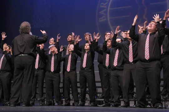 Een groep mannen, in zwart gekleed met gekleurde dassen, zingen in koor. 