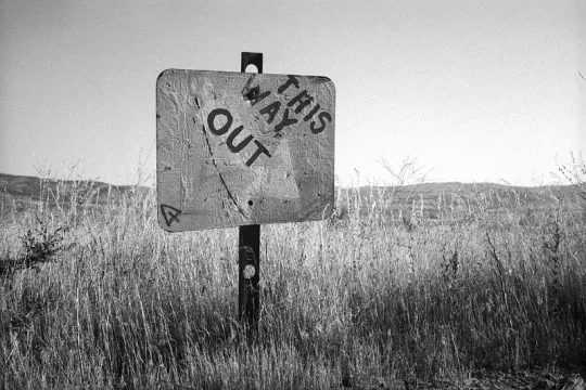Zwartwit beeld van een overschilderd bord in een dor graslandschap, met daarop de tekst 'This way out'. 