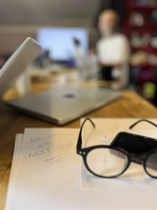 Op een blad papier, met daarop ook een opengeklapte leesbril, staat in balpen geschreven 'Apache goes Nextcloud now'. In de wazige achtergrond is een opengeklapte laptop en een man te zien.