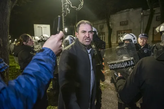 Een man kijkt boos in de camera, terwijl politie-agenten met helm en schild de straat achter hem in de gaten houden.