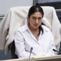 Zuhal Demir, in wit hemd, zit met een verveelde uitdrukking in het Vlaams Parlement, een witte vest over de rugleuning van haar stoel.