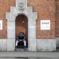 Een persoon zit op een bankje tegen de muur van een opvangcentrum van Fedasil en kijkt naar de grond. 