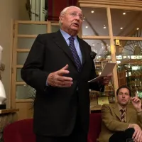 Rudolf Vanmoerkerke staat recht en leest op van een papier, geflankeerd door een journalist van Knack en een buste van een naakte vrouw. 