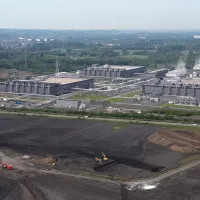 Dronebeelden tonen een industrieel gebouwencomplex aan de rand van een veld vol zand en bouwvoertuigen.